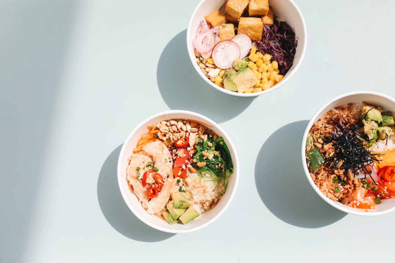 A top view of three vibrant poke bowls with varied fresh ingredients on a light blue surface.