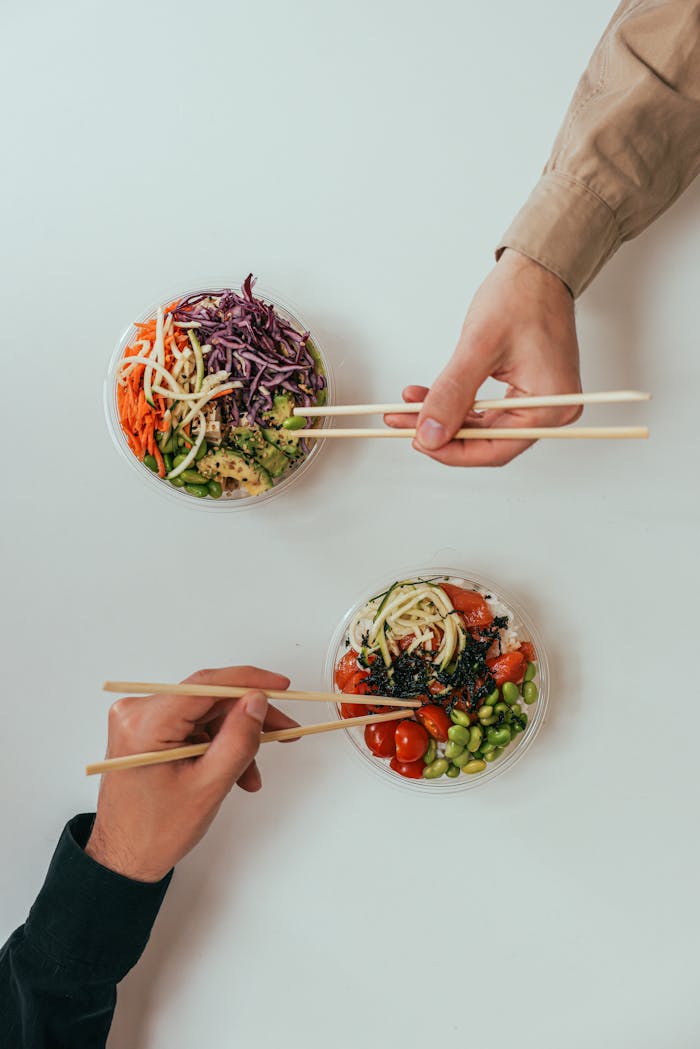 who-we-are Two people using chopsticks to enjoy fresh vegetable bowls on a white table.